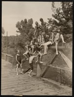 Lightly sepia-toned photograph of eleven young boys, wet from swimming, in bathing suits climbing a wooden ledge on a bridge over a creek. In the background, the creek flows underneath the bridge, surrounded by trees.